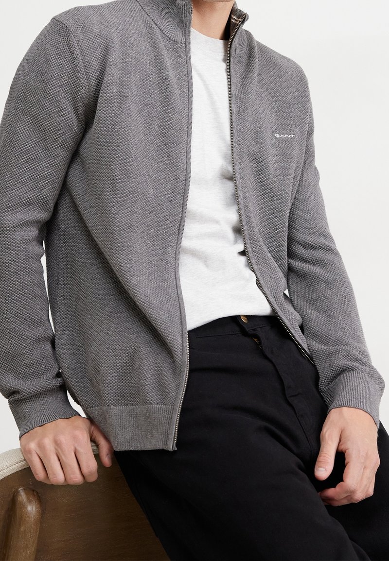 Homme portant un cardigan zippé gris texturé par-dessus un T-shirt blanc et un pantalon noir, assis sur une chaise en bois.