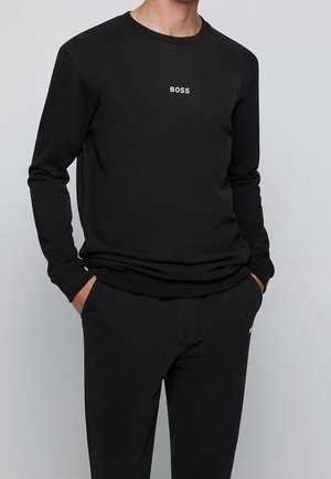 Mężczyzna w czarnej koszuli z długim rękawem z napisem "BOSS" na środku klatki piersiowej, w połączeniu z czarnymi spodniami, ręce w kieszeniach.