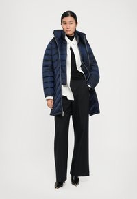 Vrouw in een marineblauwe pufferjack over een witte blouse en een zwarte coltrui, met zwarte wijde broek en zwarte puntige schoenen.