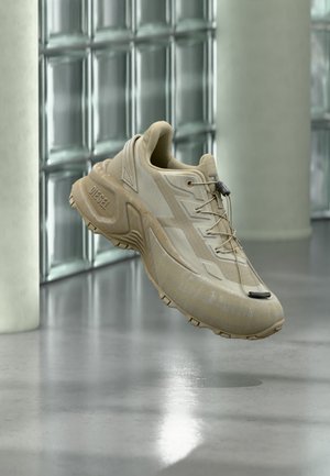 Zapatilla beige Diesel con suela gruesa y sistema de cordones con presilla flotando en interiores contra una pared de bloques de vidrio y suelo pulido.