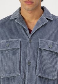 Camisa de pana en azul claro que presenta una superficie texturizada, dos bolsillos en el pecho, cierres de botón y un cuello puntiagudo.