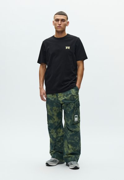 Μαύρο βαμβακερό μπλουζάκι με κίτρινο λογότυπο, σε συνδυασμό με πράσινο παντελόνι cargo με σχέδιο tie-dye, πλαϊνές τσέπες και ετικέτα, συνδυασμένο με γκρι αθλητικά παπούτσια.