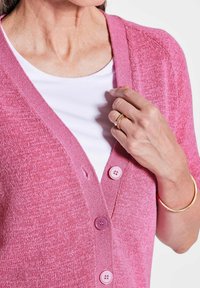 Rosa Cardigan mit V-Ausschnitt, kurzen Ärmeln und drei lila Knöpfen. Der Stoff ist strukturiert und leicht, darüber getragen wird ein weißes Oberteil.