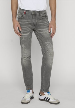 Uomo che indossa jeans grigi slavati e consumati e scarpe da ginnastica bianche con strisce nere, in piedi contro uno sfondo chiaro e semplice.