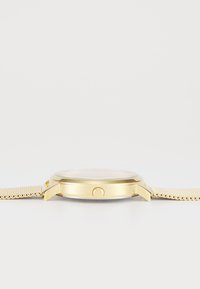 Reloj de metal dorado con una correa de malla texturizada, caja redonda y una corona simple. La esfera presenta una cubierta de vidrio liso y un diseño minimalista.
