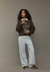 Felpa marrone con colletto bianco e logo, abbinata a pantaloni della tuta grigi. La persona tiene un dispositivo con cuffie, in piedi contro uno sfondo neutro.