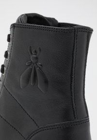 Czarny skórzany but z wytłoczonym logo owada na boku, widocznym przeszyciem i haczykami na sznurowadła.