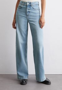 Ljusblå högmidjade vida jeans med en urtvättrad finish, fem fickor och en klassisk knapp- och dragkedja, bärs med svarta skor.