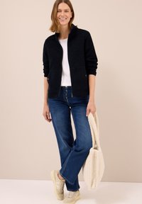 Granatowa polarowa kurtka z wysokim kołnierzem, dwoma bocznymi kieszeniami, w połączeniu z białą koszulką, niebieskimi dżinsami i jasnymi sneakersami.