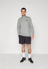 Šedý pulovr s polovičním zipem a logem, nošený přes černé šortky. Model má na sobě černé tenisky a bílé ponožky, představující atletické oblečení.