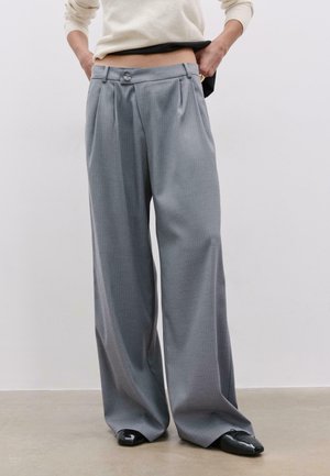 Persona che indossa pantaloni grigi a vita alta e gambe larghe con pieghe frontali, scarpe nere con punta appuntita e un maglione crema a maniche lunghe sollevando leggermente l'orlo.