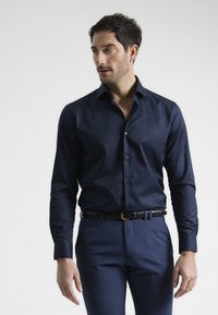Camisa de botones azul marino en tejido suave, con un cuello clásico y botones de contraste, combinada con pantalones oscuros ajustados y un cinturón negro.