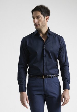 Camisa de botones azul marino en tejido suave, con un cuello clásico y botones de contraste, combinada con pantalones oscuros ajustados y un cinturón negro.