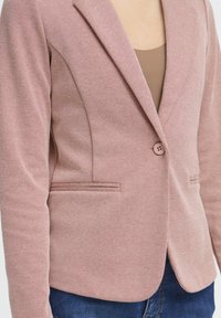 Blazer rosa empolvado hecho de tela texturizada. Cuenta con cierre de un solo botón, solapas con muesca y bolsillos laterales. Se lleva sobre una camiseta marrón.