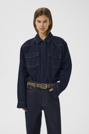 Dark denim shirt with a collar, two chest pockets, and long sleeves. Paired with high-waisted dark denim pants and a khaki belt.
