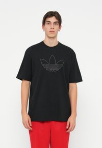 Melna kokvilnas T-krekls ar īsām piedurknēm, centrā ar punktu grafiku Adidas trīs lapu logo. Valkāts ar sarkaniem biksēm.