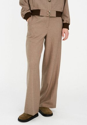 Pantalones beige de pierna ancha hechos de tela texturizada, con frente plano, bolsillos laterales y corte recto. Acompañados de calzado marrón.
