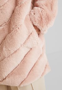 Chaqueta de pelo sintético de color rosa claro con un patrón de chevron, que presenta una textura suave y un puño de manga ajustado. Detalles de costura visibles en la tela.