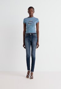 T-shirt aderente blu con grafica di un delfino, abbinato a jeans skinny scuri e sandali con tacco argento. Tessuto morbido, stile casual.
