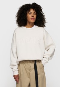 Felpa beige chiaro con lettering in rilievo "10DAYS", maniche larghe e una vestibilità rilassata. Abbinata a pantaloni chiari.
