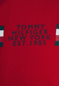Červené bavlněné tričko s ribkovanou texturou, se tmavě modrým nápisem: "TOMMY HILFIGER NEW YORK EST. 1985" a malými barevnými blokovými akcenty.