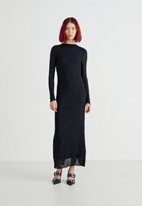 Dolga črna obleka z teksturirano blago, dolgimi rokavi in ravno silhueto. Ujemajoče se s črnimi čevlji s studnimi. Preprost in eleganten dizajn.