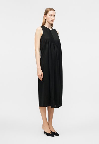 Γυναίκα που στέκεται, φορώντας μαύρο μίντι φόρεμα χωρίς μανίκια με πιετέ λεπτομέρεια μπροστά και μαύρα μυτερά παπούτσια με χαμηλό τακούνι, μπροστά σε απλό ανοιχτό γκρι φόντο.