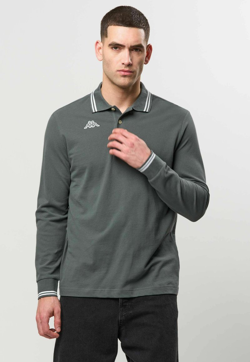 Homme aux cheveux courts et foncés portant un polo à manches longues gris foncé avec un col et des poignets à rayures blanches, debout devant un fond uni.