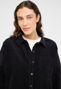 Camisa de pana negra con cierre de botones en la parte delantera, cuello y un bolsillo en el lado izquierdo del pecho; textura suave y patrón estriado.