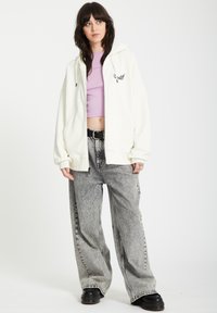 Felpa bianca con zip, dotata di ampie tasche frontali e cappuccio regolabile, abbinata a jeans larghi con motivo bianco e nero e stivali neri chunky.