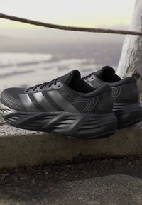 Pair of black running shoes with textured soles and striped design placed on a stone surface outdoors near a rope barrier.