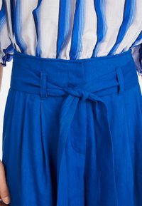 Blauer Leinenbund mit einem gebundenen Knoten, ausgestattet mit Falten und einer Seitentasche. Der Stoff hat eine glatte Textur und eine lebendige Farbe.