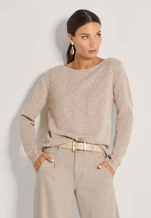 Femme portant un pull en maille torsadée beige, un pantalon beige avec une ceinture assortie, et des boucles d'oreilles pendantes en perles, debout les mains dans les poches regardant vers la droite.