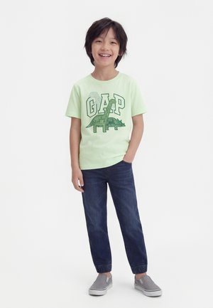 Αγόρι που χαμογελάει φορώντας ένα ανοιχτοπράσινο μπλουζάκι GAP με γραφικό δεινόσαυρου, σκούρο μπλε τζιν και γκρι παπούτσια χωρίς κορδόνια, στέκεται με το ένα χέρι στην τσέπη.