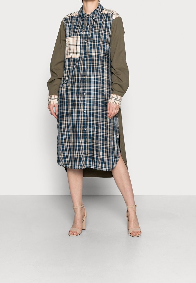 Kariertes Hemdkleid in Marineblau und Beige mit durchgehender Knopfleiste, langen Ärmeln und asymmetrischem Saum. Mit einer Brusttasche und seitlichen Schlitzen.