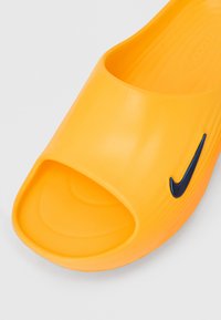 Gul åben tå-slide sandal med sort Nike swoosh-logo på siden og tekstureret fodseng på en hvid baggrund.
