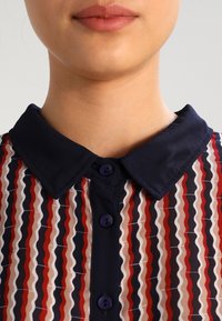 Camicia colletto con un motivo a strisce ondulate in blu navy, rosso e beige. Presenta bottoni viola e una texture del tessuto liscia.