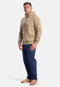 Beige hoodie, katoenmix, voorzak met kangoeroe-stijl, marineblauwe broek, beige sandalen. Model staat met een neutrale uitdrukking.