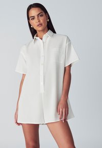 Bela srajca s kratkimi rokavi, z gumbno zaponko spredaj, enim žepom na prsih in sproščenim krojem, izdelana iz teksturirane tkanine z gladkim zaključkom.