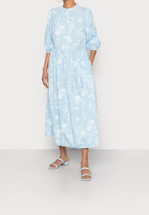 Vrouw draagt een lichtblauwe midi-jurk met witte bloemenpatronen, pofdriekwart mouwen en bijpassende lichtblauwe platte sandalen.