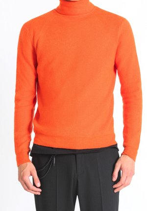 Mężczyzna ubrany w jaskrawo pomarańczowy golf w połączeniu z czarnymi spodniami w drobną prążek oraz czarną łańcuszkową ozdobą po lewej stronie.