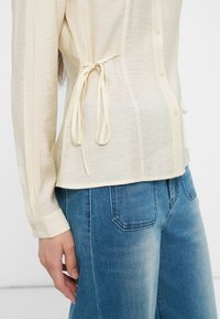 Μπεζ μπλούζα με μακριά μανίκια και δεσίματα στο πλάι, φορεμένη με ξεθωριασμένο μπλε τζιν, με τον καρπό και το αριστερό χέρι χαλαρά στο πλάι.