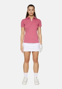 Roze polo met kraag en knopenlijst, gecombineerd met een witte skort. Witte sportieve schoenen en een lichte handschoen maken de outfit af.