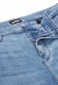 Jeans in denim azzurro chiaro con zip frontale, chiusura con bottone in metallo e etichetta con marchio. Il tessuto presenta una finitura sfumata e strutturata.
