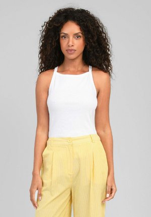 Femme aux cheveux bouclés porte un haut blanc sans manches et un pantalon jaune rayé taille haute, debout devant un fond gris uni.