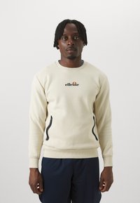Kremeni dukserica s malim crnim "ellesse" logotipom, s crnim patentnim zatvaračima na stranama i rebrastim manžetama. Nosena s tamnoplave hlače.