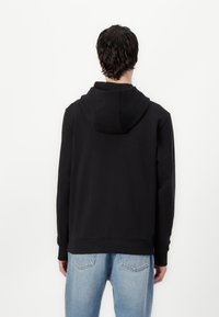 Zwarte hoodie van een katoenmix, met een trekkoordkap, ribbels aan de mouwen en onderkant. De achterkant is eenvoudig en heeft geen bedrukkingen. Gedragen met lichtblauwe spijkerbroek.