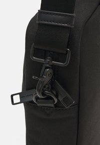 Svart veske med teksturert stoff, med avtakbar stropp og metallelementer inkludert en klips og logo-tags.