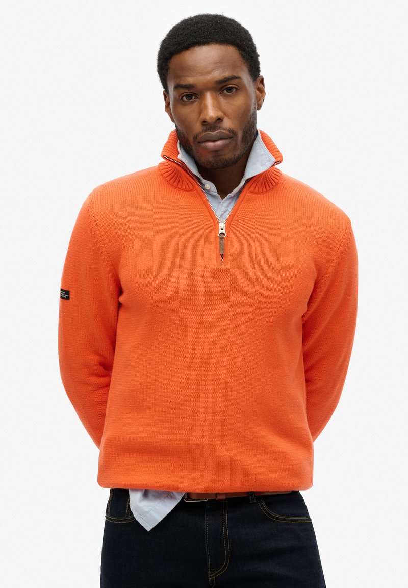 Oranger Strickpullover mit hohem Kragen und Reißverschluss, aus strukturiertem Material und mit einer kontrastierenden hellblauen Bluse darunter.