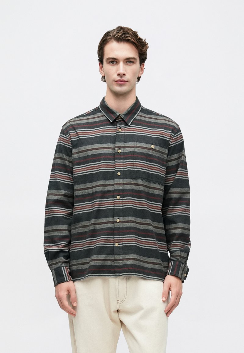 Młody mężczyzna w koszuli z długim rękawem zapinanej na guziki w poziome szare, czarne i czerwone paski, w połączeniu z off-white spodniami, zwrócony w stronę z przodu.